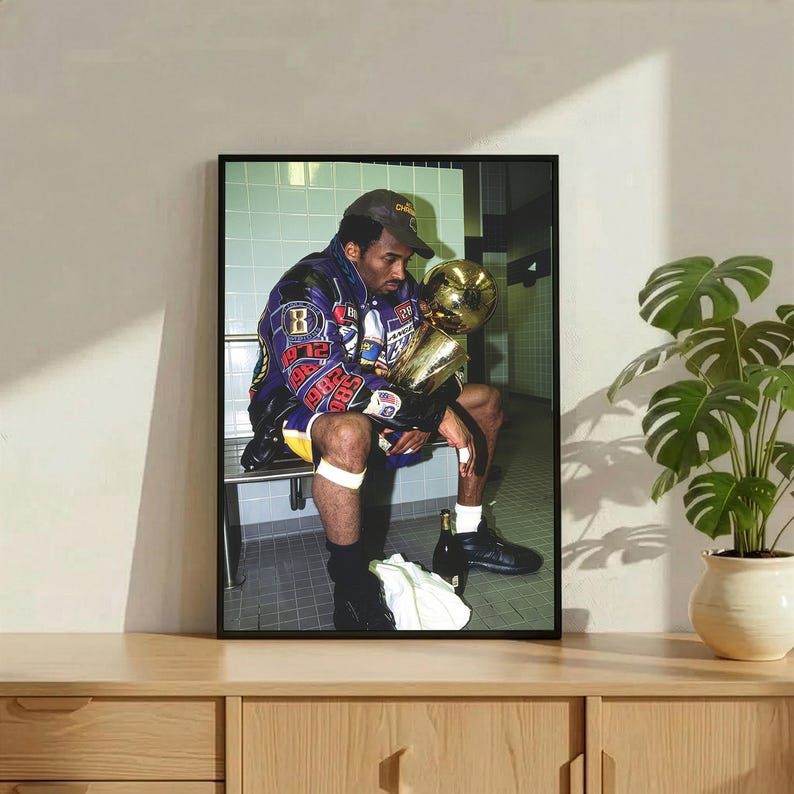 May include: Framed print featuring a seated figure in a purple and gold jacket, holding a gold trophy. The image is set against a tiled wall background. The figure is wearing a baseball cap and shorts.