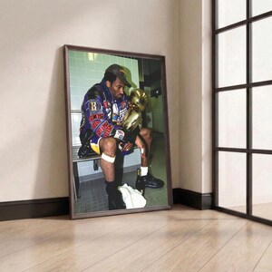 May include: Framed print featuring a seated figure holding a gold trophy. The figure is wearing a purple and blue jacket, a baseball cap, and shorts. The background shows a tiled wall and a bench. The print is leaning against a wall.