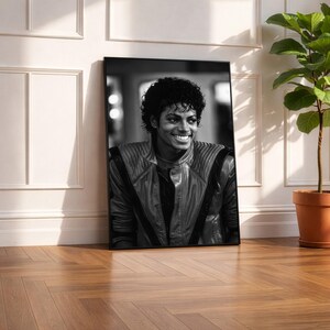 May include: Black and white framed portrait of a smiling man with curly hair, wearing a leather jacket. The portrait is leaning against a white wall, next to a potted plant on a wooden floor.