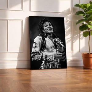 May include: Black and white framed print of a smiling man holding a microphone. The man is wearing a light-colored shirt with a black harness and a decorative belt. The print is leaning against a white wall and a wooden floor.