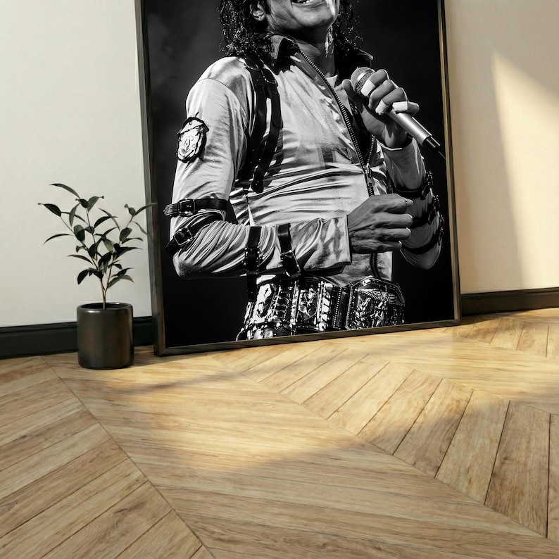 May include: Black and white framed print of a performer holding a microphone. The performer is wearing a light-colored satin shirt with a black harness and belt. The image is leaning against a wall, with a small potted plant in the corner.