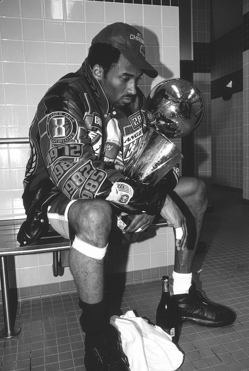 May include: Black and white image of a person wearing a baseball cap and a jacket with patches and the word "Champions" on the cap. The person is holding a trophy and a globe. There are two bottles and a towel on the floor.