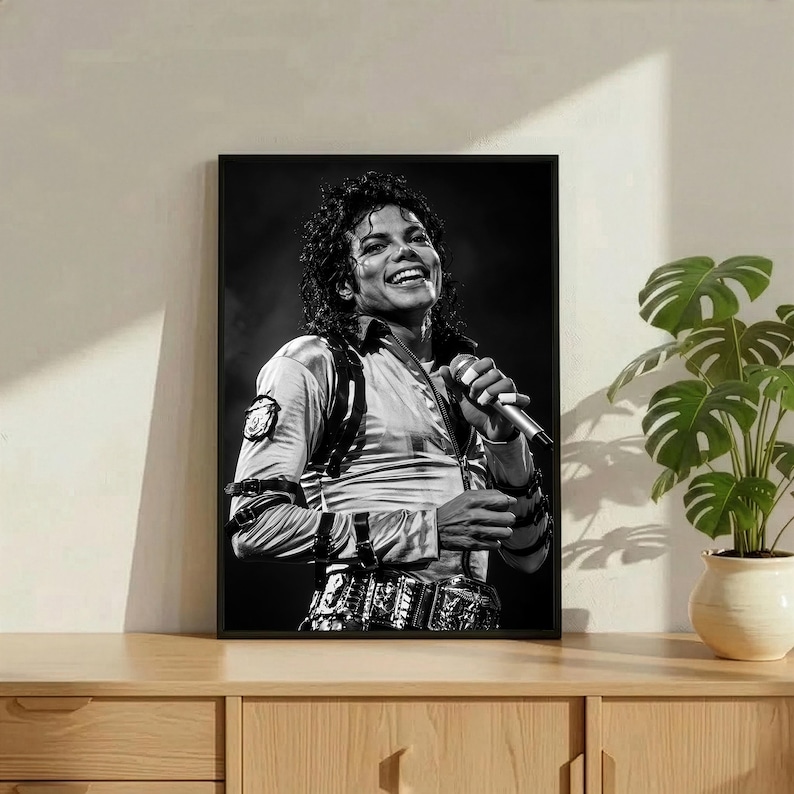 May include: Black and white framed print of a smiling person holding a microphone. The person is wearing a light-colored shirt, a belt with decorative details, and has curly hair. The print is displayed on a wooden cabinet.