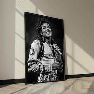 May include: Black and white framed print of Michael Jackson smiling while holding a microphone. He is wearing a light-colored satin shirt with a patch on the left shoulder, and a decorative belt. The image is leaning against a wall.