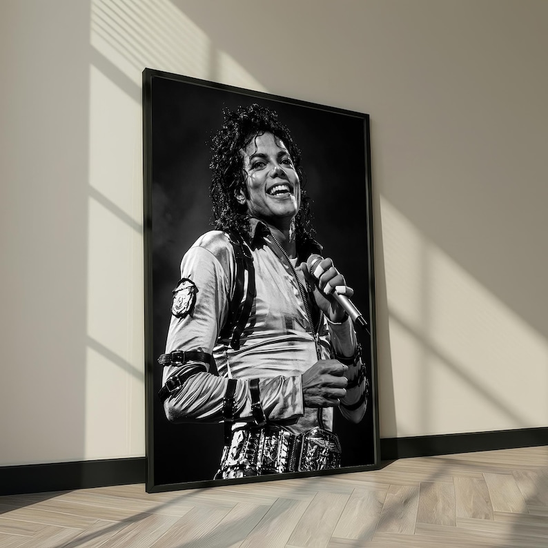 May include: Black and white framed print of Michael Jackson smiling while holding a microphone. He is wearing a light-colored satin shirt with a patch on the left shoulder, and a decorative belt. The image is leaning against a wall.