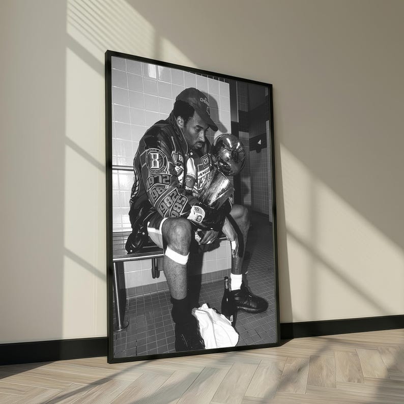 May include: Black and white framed print of a person sitting on a bench, holding a trophy. The person is wearing a baseball cap, a leather jacket with patches, and athletic shorts. The image is in a tiled room.