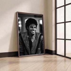 May include: A framed black and white portrait of Michael Jackson, leaning against a wall. The portrait features a smiling Michael Jackson wearing a leather jacket. The frame is brown, and the background includes a window and wooden floor.