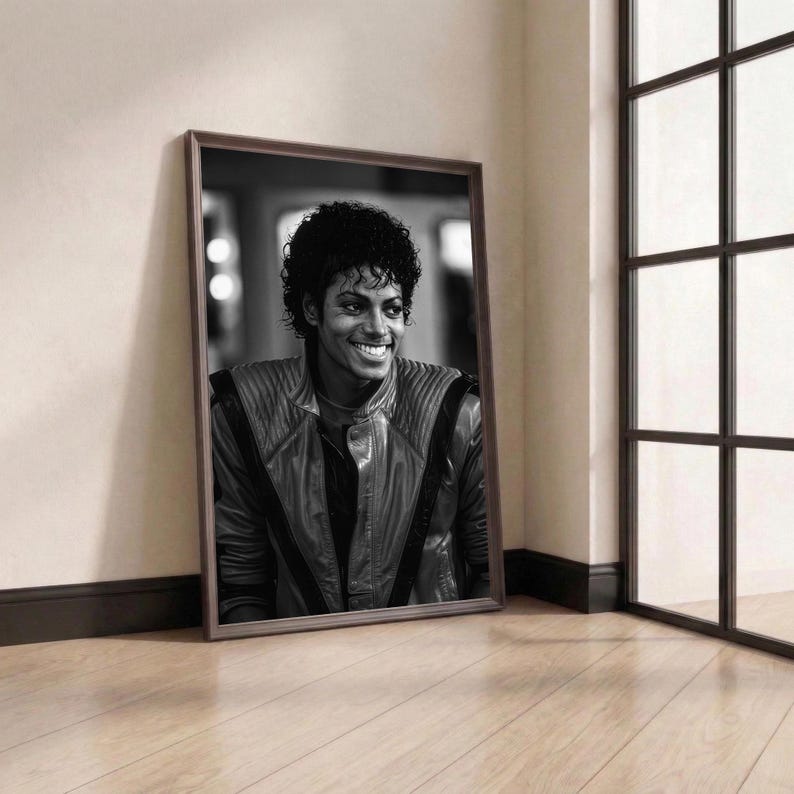 May include: A framed black and white portrait of Michael Jackson, leaning against a wall. The portrait features a smiling Michael Jackson wearing a leather jacket. The frame is brown, and the background includes a window and wooden floor.