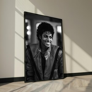May include: Black and white framed portrait of a smiling person with curly hair. The person is wearing a leather jacket with a high collar. The portrait is leaning against a wall.