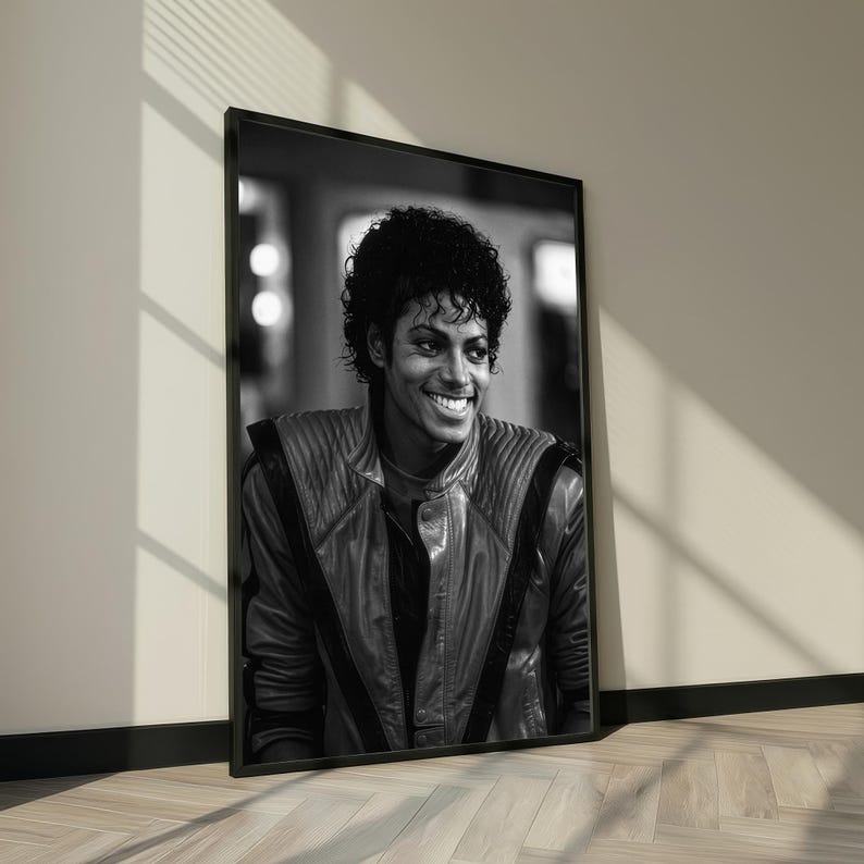 May include: Black and white framed portrait of a smiling person with curly hair. The person is wearing a leather jacket with a high collar. The portrait is leaning against a wall.