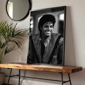May include: Black and white framed portrait of Michael Jackson smiling. He wears a leather jacket with quilted shoulders. The portrait is displayed on a wooden bench with black metal legs, near a plant and a round mirror.