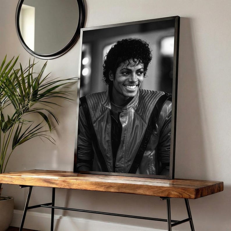 May include: Black and white framed portrait of Michael Jackson smiling. He wears a leather jacket with quilted shoulders. The portrait is displayed on a wooden bench with black metal legs, near a plant and a round mirror.