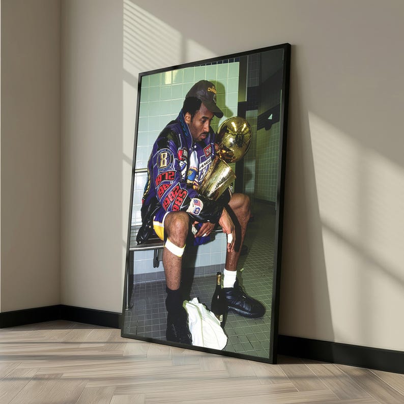 May include: Framed print featuring a person seated, holding a gold trophy. The person wears a purple and gold jacket and a baseball cap. The image is set in a locker room setting, with a black frame.