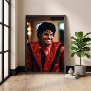 May include: Framed portrait of a smiling person wearing a red leather jacket with black stripes. The portrait is displayed on a wall next to a window and a potted plant. The room has wooden floors.