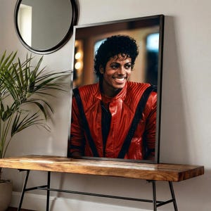 May include: Framed portrait of a smiling person wearing a red leather jacket with black accents. The portrait is displayed on a wooden bench with black metal legs, with a round mirror and a potted plant in the background.