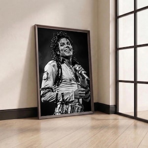 May include: Framed black and white portrait of Michael Jackson, smiling and holding a microphone. The image is in a brown frame, leaning against a wall. The room has a window with black frames and a wooden floor.