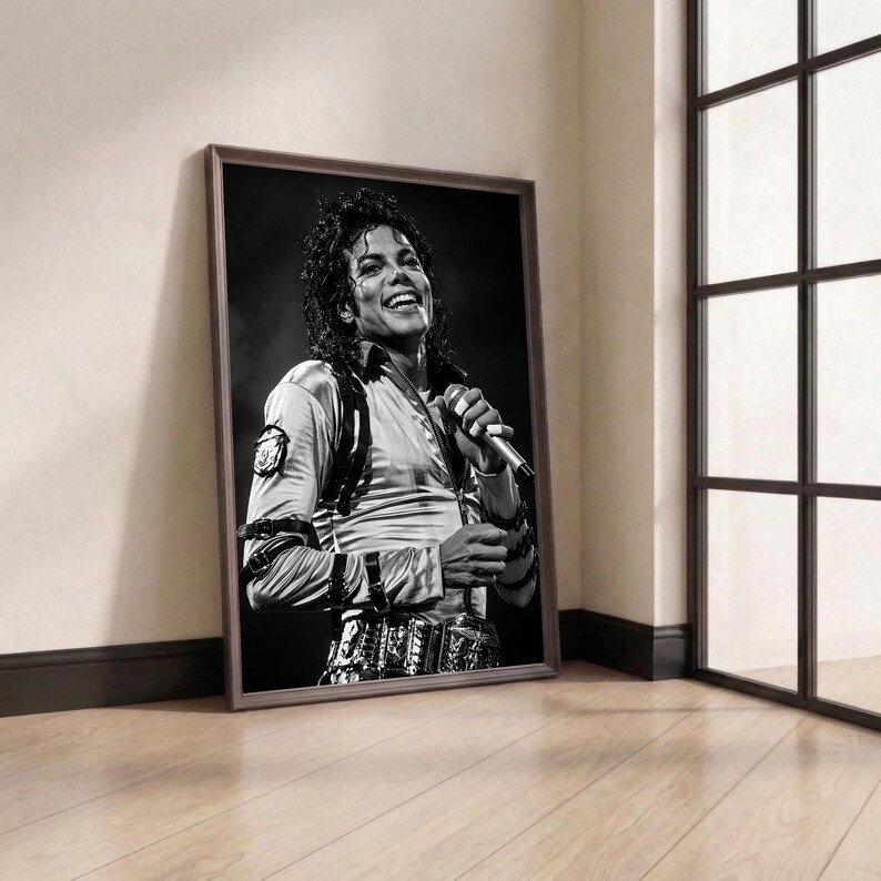 May include: Framed black and white portrait of Michael Jackson, smiling and holding a microphone. The image is in a brown frame, leaning against a wall. The room has a window with black frames and a wooden floor.