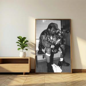 May include: Black and white framed print of a man in a leather jacket and baseball cap, holding a trophy. The man is seated on a bench, with a towel and bottles nearby. The image is in a room with a wooden floor.