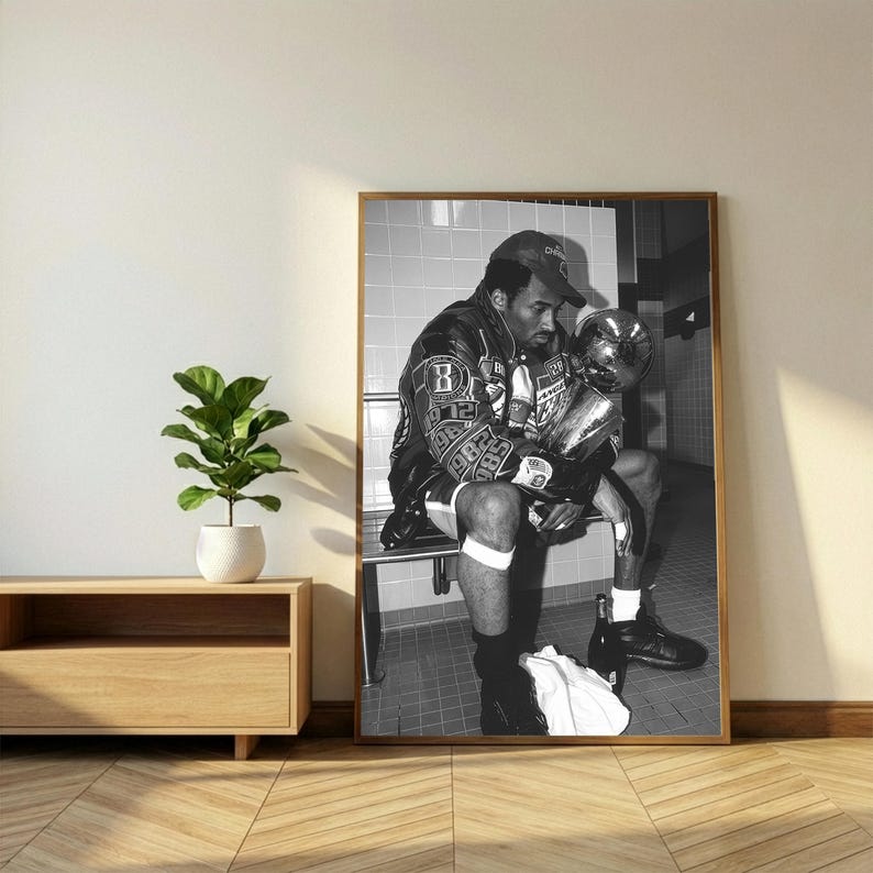 May include: Black and white framed print of a man in a leather jacket and baseball cap, holding a trophy. The man is seated on a bench, with a towel and bottles nearby. The image is in a room with a wooden floor.