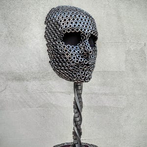 Op de afbeelding: Een metalen sculptuur van een gezicht gemaakt van moeren en bouten. Het gezicht is gemonteerd op een gedraaide metalen standaard met een ronde basis.