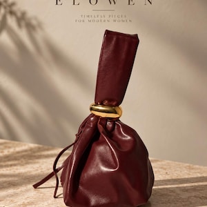 May include: A deep burgundy leather pouch bag featuring a smooth, rounded gold-tone metal ring accent at the top handle. The bag has a gathered, soft structure with a drawstring closure. The text ELOWEN TIMELESS PIECES FOR MODERN WOMEN is visible.