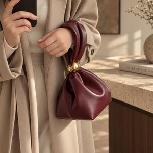 May include: A deep burgundy pouch handbag featuring a soft, gathered silhouette and a single knotted handle. The handle is accented with a large, polished gold-tone spherical bead at the base, providing a metallic contrast to the smooth, dark red material.