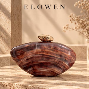 Puede incluir: Un bolso de mano tipo clutch de forma ovalada con un acabado de resina veteada en tonos marrón y crema. El bolso presenta una silueta lisa y redondeada con un cierre metálico dorado en la parte superior. El texto ELOWEN aparece impreso en negro sobre el bolso.