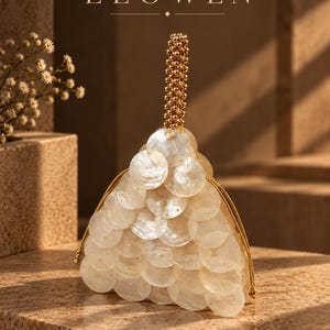 Peut inclure: Un sac à main triangulaire recouvert de disques de coquillages blancs irisés superposés. Le sac présente une poignée supérieure en perles dorées et des cordons de serrage dorés. Le mot ELOWEN est imprimé en blanc au-dessus du sac.
