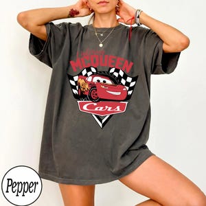 May include: Dark gray t-shirt featuring a graphic of Lightning McQueen from the movie Cars. The design includes the text "Lightning McQueen" in red, the word "Cars" in white, and a checkered flag.