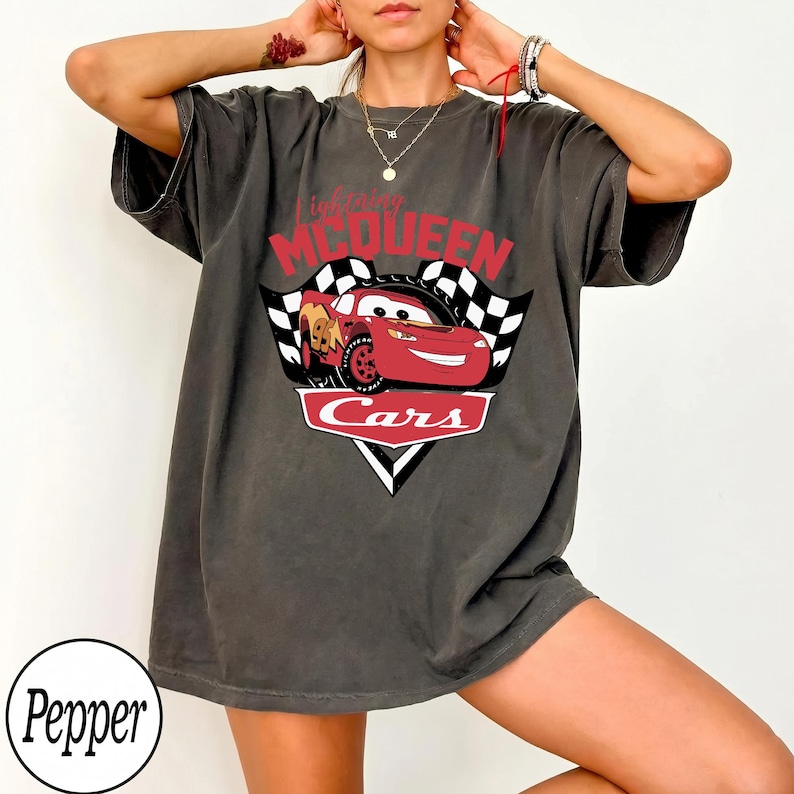 May include: Dark gray t-shirt featuring a graphic of Lightning McQueen from the movie Cars. The design includes the text "Lightning McQueen" in red, the word "Cars" in white, and a checkered flag.
