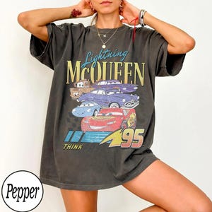 Puede incluir: Camiseta gris oscuro con un gr&aacute;fico de Rayo McQueen y otros personajes de Cars. El dise&ntilde;o incluye el texto "Lightning McQueen" y "95". La camiseta tiene un aspecto vintage y desgastado.