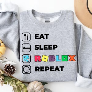 Puede incluir: Una sudadera gris jaspeado de cuello redondo con un estampado gr&aacute;fico de cuatro iconos y texto. El texto dice EAT, SLEEP, ROBLOX y REPEAT en negro, con la palabra ROBLOX mostrada en letras de colores junto a su icono correspondiente.