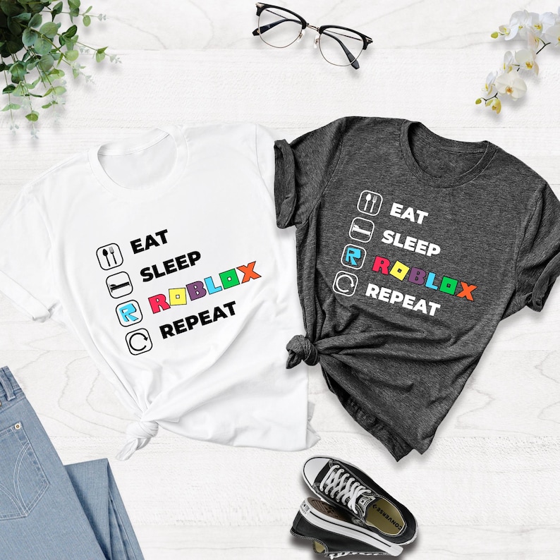 Puede incluir: Dos camisetas de manga corta, una blanca y otra gris oscuro, cada una con un estampado gr&aacute;fico con el texto EAT, SLEEP, ROBLOX, REPEAT. El dise&ntilde;o incluye iconos de comer, dormir y repetir, con la palabra ROBLOX en letras de colores.