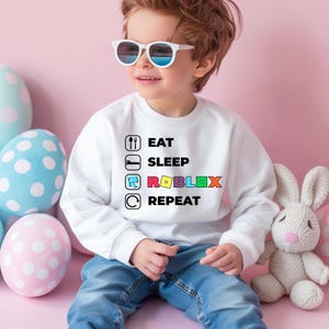 Puede incluir: Una sudadera blanca de manga larga con un estampado gr&aacute;fico que incluye el texto EAT SLEEP ROBLOX REPEAT junto a iconos de un tenedor y una cuchara, una cama, el logotipo de Roblox y una flecha circular. Pantalones vaqueros azules.