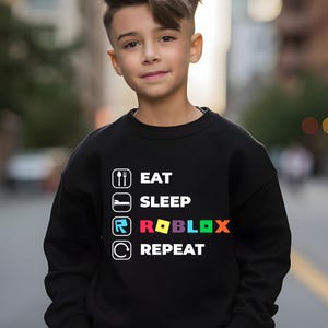 Puede incluir: Una sudadera negra de manga larga con un estampado gr&aacute;fico blanco con el texto EAT SLEEP ROBLOX REPEAT. El dise&ntilde;o incluye iconos de cuchara y tenedor, una cama, el logotipo de Roblox y una flecha circular, con la palabra ROBLOX en letras de colores.