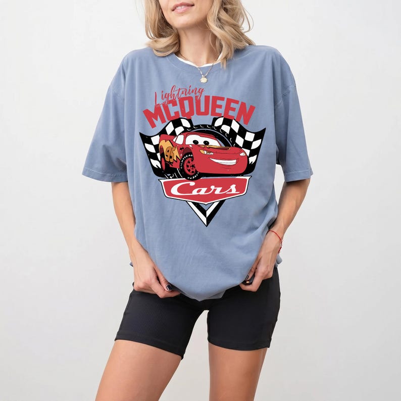 May include: A light blue t-shirt featuring a graphic of Lightning McQueen from the Cars movie. The design includes the text "Lightning McQueen" in red script above the car, with "Cars" in a white font below. The shirt has short sleeves.