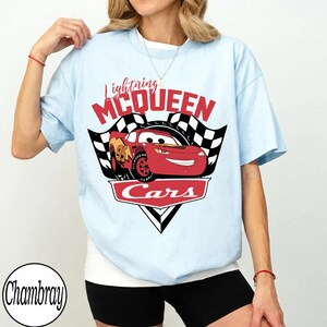 May include: Light blue t-shirt featuring a graphic of Lightning McQueen from the movie Cars. The design includes the text "Lightning McQueen" in red, a checkered flag, and the word "Cars".