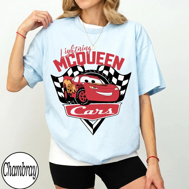 May include: Light blue t-shirt featuring a graphic of Lightning McQueen from the movie Cars. The design includes the text "Lightning McQueen" in red, a checkered flag, and the word "Cars".