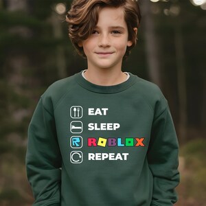 Puede incluir: Una sudadera de cuello redondo de color verde oscuro con un estampado gr&aacute;fico blanco con el texto EAT SLEEP ROBLOX REPEAT. El dise&ntilde;o incluye iconos de cuchara y tenedor, una cama, el logotipo de Roblox y una flecha circular, con la palabra ROBLOX en letras de colores.
