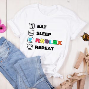Puede incluir: Una camiseta blanca de manga corta con un estampado gr&aacute;fico que incluye cuatro iconos circulares y texto. El texto dice EAT, SLEEP, ROBLOX y REPEAT, con la palabra ROBLOX en letras de colores junto a los iconos correspondientes.