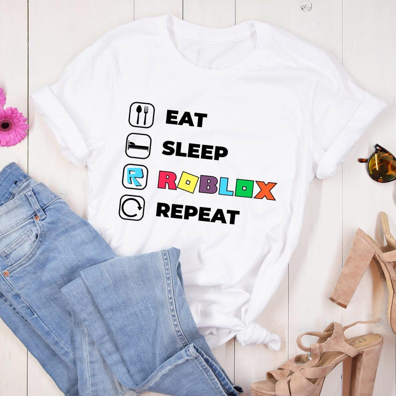 Puede incluir: Una camiseta blanca de manga corta con un estampado gr&aacute;fico que incluye cuatro iconos circulares y texto. El texto dice EAT, SLEEP, ROBLOX y REPEAT, con la palabra ROBLOX en letras de colores junto a los iconos correspondientes.