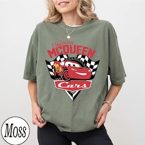 May include: Olive green t-shirt featuring a graphic of Lightning McQueen from the Cars movie. The design includes the text "Lightning McQueen" in red, a red race car, and the word "Cars" in a white outlined red banner.