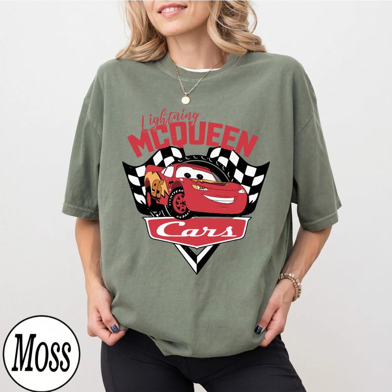 May include: Olive green t-shirt featuring a graphic of Lightning McQueen from the Cars movie. The design includes the text "Lightning McQueen" in red, a red race car, and the word "Cars" in a white outlined red banner.