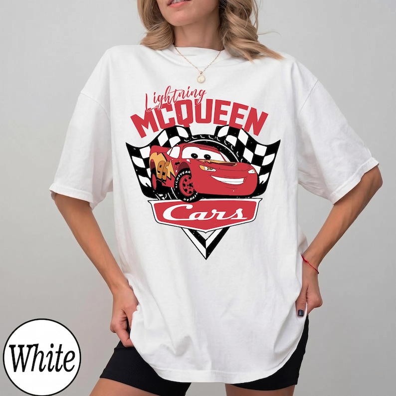 May include: White t-shirt featuring a graphic of Lightning McQueen from the Cars movie. The design includes the text "Lightning McQueen" above a red race car, with checkered flags and the word "Cars" below.
