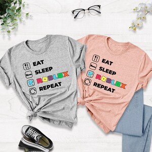 Puede incluir: Una camiseta gris jaspeado y una camiseta color melocot&oacute;n, cada una con un estampado gr&aacute;fico negro que dice EAT SLEEP ROBLOX REPEAT. El texto incluye iconos de tenedor y cuchara, una cama, la letra R y una flecha circular, con ROBLOX en letras de colores.