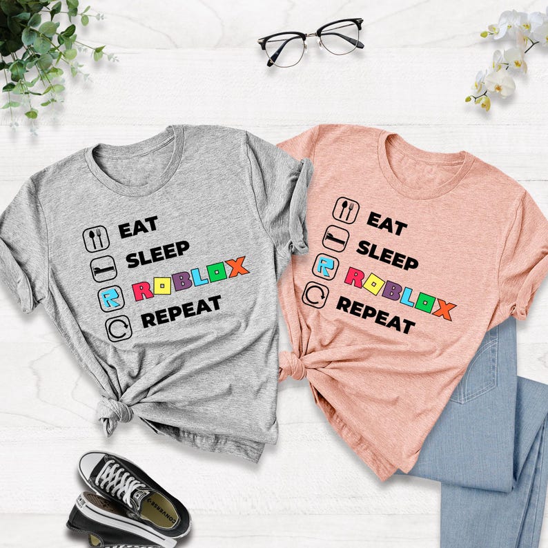 Puede incluir: Una camiseta gris jaspeado y una camiseta color melocot&oacute;n, cada una con un estampado gr&aacute;fico negro que dice EAT SLEEP ROBLOX REPEAT. El texto incluye iconos de tenedor y cuchara, una cama, la letra R y una flecha circular, con ROBLOX en letras de colores.