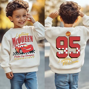 May include: Cream-colored Lightning McQueen sweatshirt with red and black graphics. The front features the text "Lightning McQueen" and a cartoon car. The back displays the number "95" and the text "Lightning McQueen".