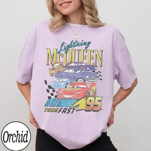 Puede incluir: Camiseta lila claro con un gr&aacute;fico de Rayo McQueen y otros personajes de Cars. El dise&ntilde;o incluye el texto "Lightning McQueen", "Think Fast" y el n&uacute;mero "95". La camiseta tiene mangas cortas.