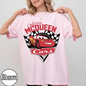May include: Light pink t-shirt featuring a graphic of Lightning McQueen from the Cars movie. The design includes the text "Lightning McQueen" in red, a checkered flag, and the word "Cars".