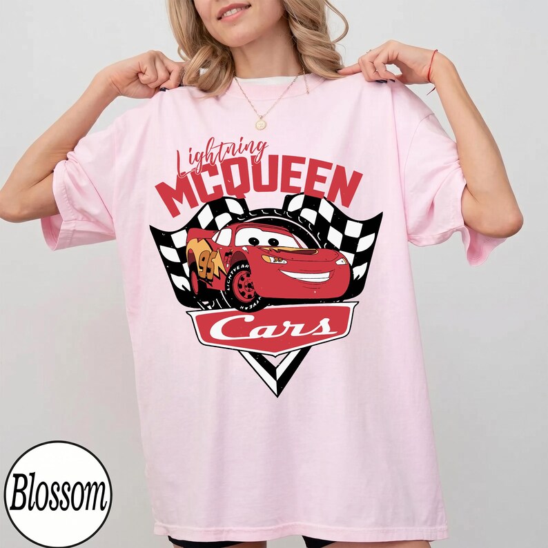 May include: Light pink t-shirt featuring a graphic of Lightning McQueen from the Cars movie. The design includes the text "Lightning McQueen" in red, a checkered flag, and the word "Cars".